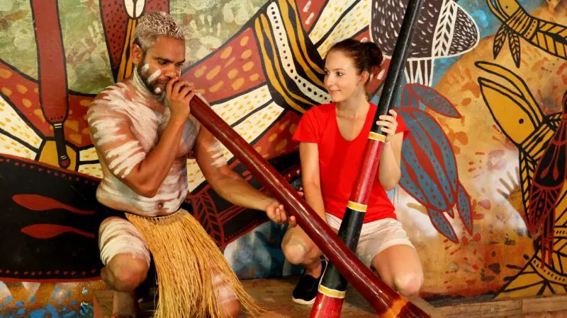 A man in traditional dress plays a didgeridoo as a woman in a red shirt observes during the renowned Kuranda Scenic Railway tour.