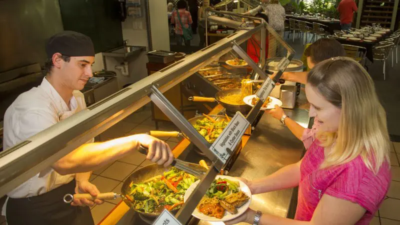 A chef serves fresh vegetables to a guest at a buffet counter while diners enjoy the renowned Classic Kuranda Scenic Railway tour.