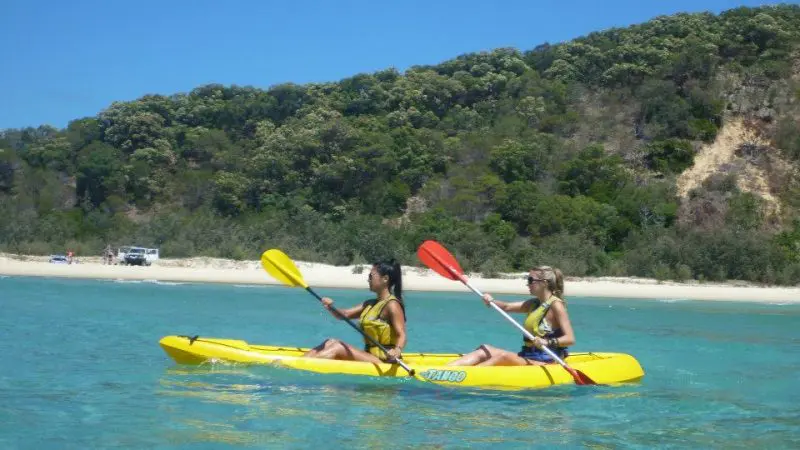 Kayakers paddle on crystal-clear blue water by Rainbow Beach’s lush green hills, enjoying a top-rated Great Beach Drive adventure.