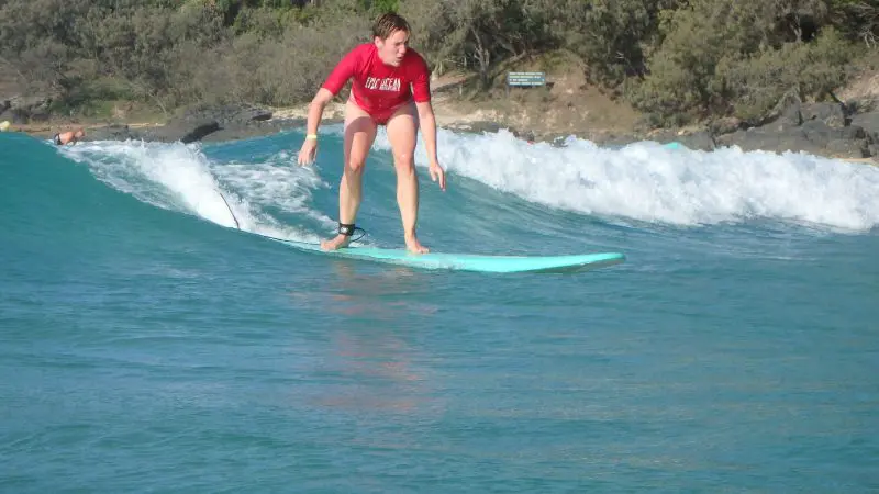 Surfer in vibrant red shirt riding Australia’s Longest Wave by a pristine, sandy, tree-lined shore under a bright blue sky.