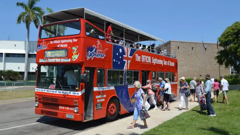 Tourists board the vibrant red Croc Explorer sightseeing bus under sunny skies by a lush green lawn, ready for an unforgettable adventure.
