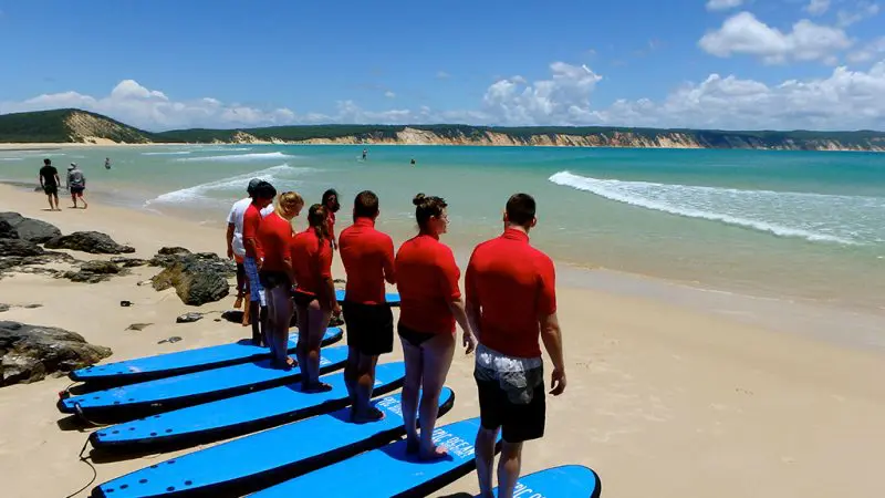 Surf lesson at Australia's Longest Wave Beach Drive: group in red shirts on blue surfboards faces sea, Learn To Surf school.