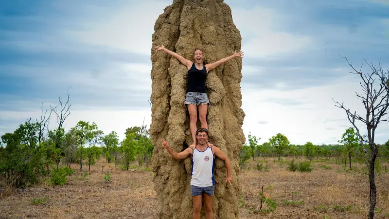 Two travellers smile, one perched atop a tall termite mound and one standing, on a 10 Day Broome to Darwin Tour adventure in Australia.