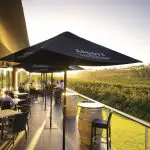 Guests enjoy al fresco dining under parasols at sunset, with vineyard views on the McLaren Vale North Hop-On Hop-Off Wine Tour.