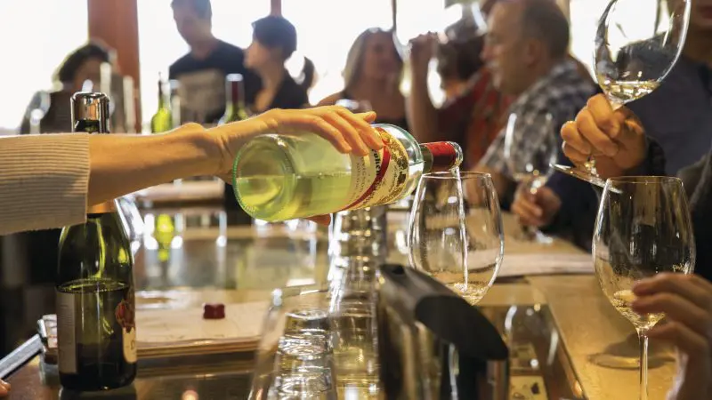 Someone pours premium white wine at a bustling McLaren Vale North Hop-On Hop-Off Tour tasting bar, enhancing wine experience.