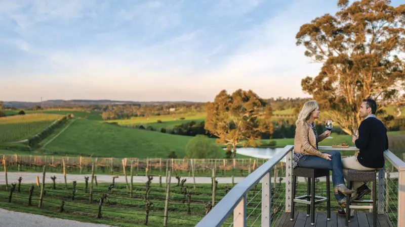 Couple savouring gourmet wine and cuisine on a scenic deck with sweeping vineyard views on a Hahndorf Adelaide Hills wine tour.