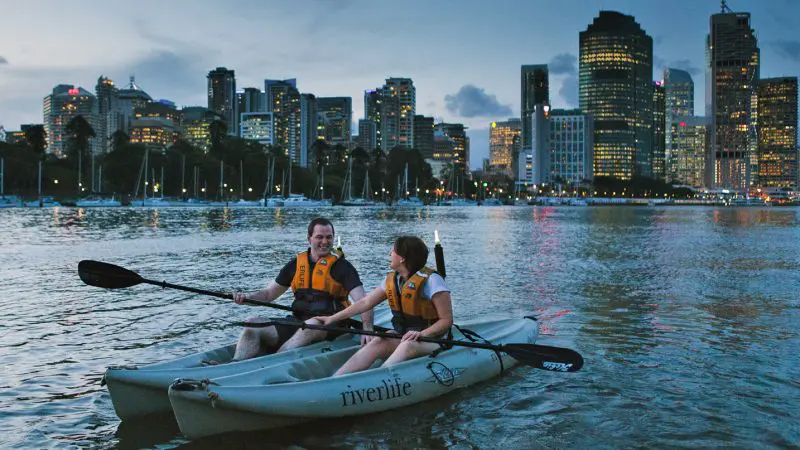 Two people kayaking on a river at sunset with city skyline and high-rise buildings in the background, enjoying a 1.5-hour kayak hire.