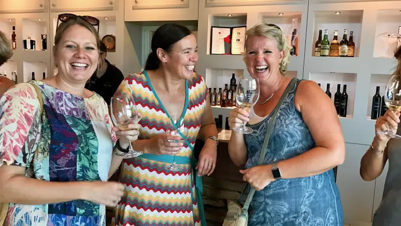 Three women enjoying a Full Day Swan Valley Wine Tour, laughing and toasting wine glasses with a backdrop of premium wine bottles.