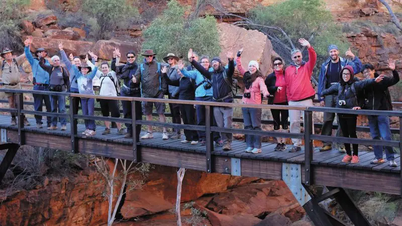 Tour group on 3 Day Uluru Rock The Centre Tour stands smiling and waving on a wooden bridge in a scenic, rugged rocky gorge.