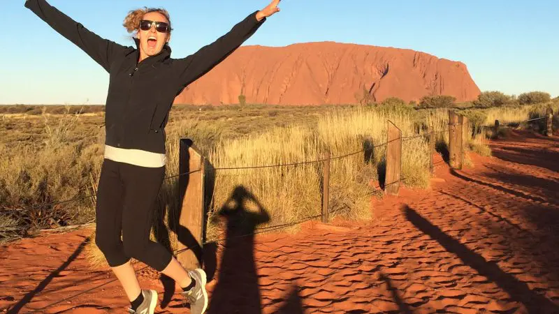 Energetic woman leaps with joy on a scenic red dirt track during the 3 Day Uluru Rock The Centre Tour from Yulara to Alice Springs.