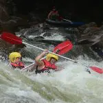 Adventurous duo in helmets kayaking fierce rapids during a Half Day Sports Rafting trip, paddles lifted in thrilling excitement.