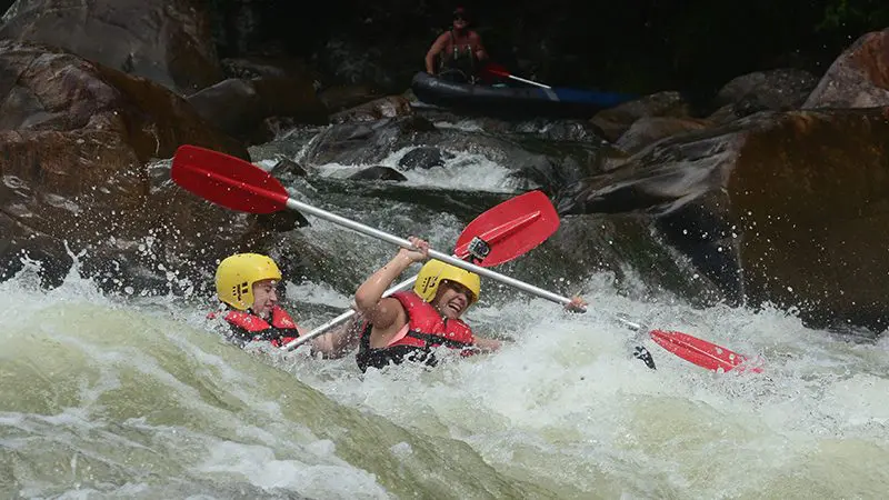 Adventurous duo in helmets kayaking fierce rapids during a Half Day Sports Rafting trip, paddles lifted in thrilling excitement.