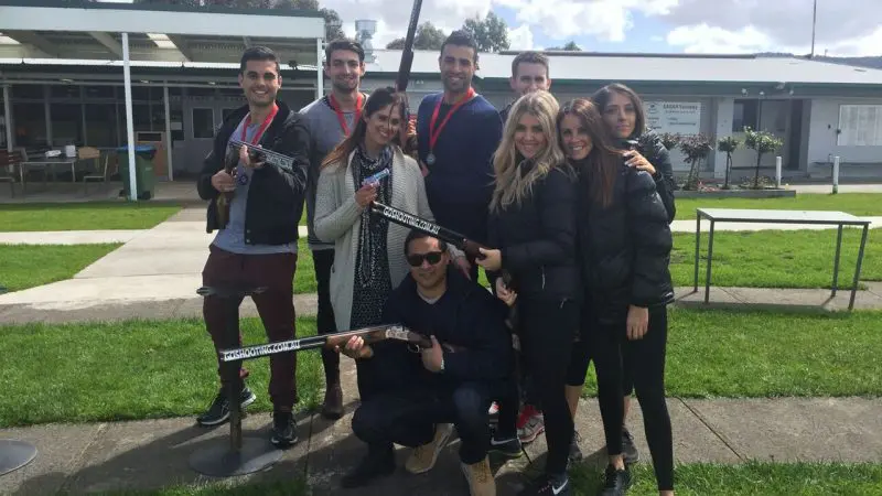 A smiling group at Vic Werribee stands outdoors on grass, proudly displaying medals and prop shotguns under a cloudy sky.