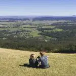 Two people relax on a lush grassy hill overlooking scenic Gold Coast valleys, envisioning a luxurious Ultimate Gourmet Tasting Experience.
