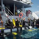 Scuba divers in PADI Open Water Course 2 sit on a boat’s edge, fully equipped with fins and kit, preparing to dive into the sea.