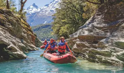 Adventurers in life jackets paddle a vibrant red raft down the crystal-clear Dart River, framed by rugged cliffs and majestic mountains.