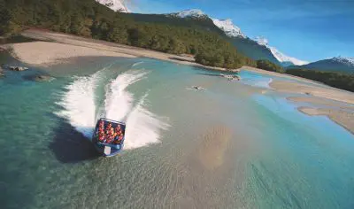 A speedboat filled with passengers speeds along the crystal-clear Dart River, leaving a white wake amid lush forests and snow-capped mountains.