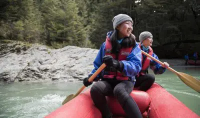 Adventurers in life jackets paddle a vibrant red Funyaks raft on the scenic Dart River, framed by lush trees and rugged rocks.