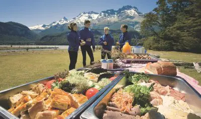 Four people enjoy a scenic Dart River Funyaks trip, standing by a picnic table with majestic mountains and lush trees in the background.