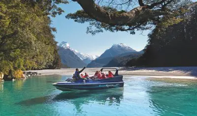 Adventurers riding a blue jet boat speed along the crystal-clear Dart River, flanked by majestic mountains, lush trees, and Funyaks.