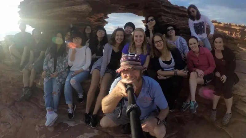 Tour group enjoys a selfie atop red rocks beneath a striking stone arch, bathed in sunshine on the 6 Day Perth to Exmouth adventure.