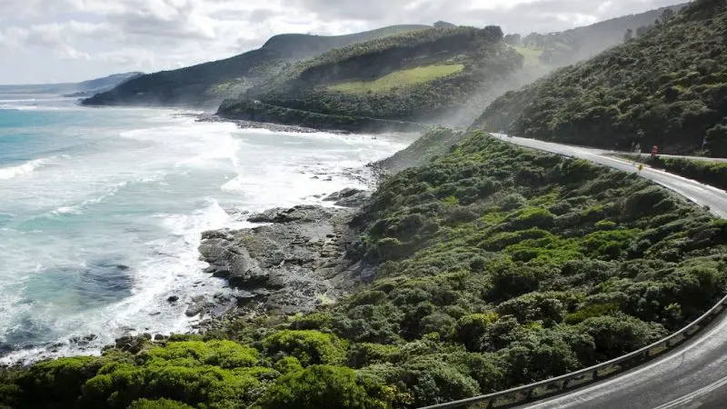 Scenic Great Ocean Road curves by lush green hills at sunset tour, with dramatic waves crashing along the picturesque Australian coastline.
