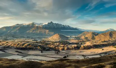 Breathtaking snow-capped peaks tower over a sunlit Queenstown valley, with lush fields and trees beneath a dramatic cloudy sky.