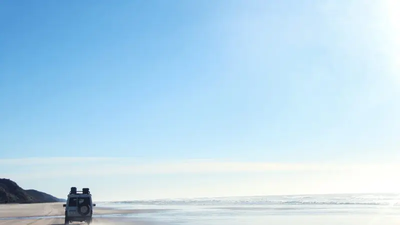 4WD vehicle drives along sandy beach during a 3 Day K'gari Adventure, with dormitory accommodation near Rainbow Beach beneath clear blue skies.