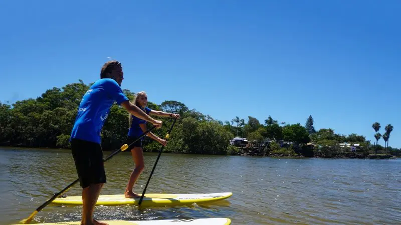 Experience a private 2-hour Byron Bay stand up paddle board nature tour; two people glide on calm waters with scenic trees and houses nearby.