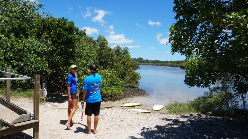 Two people enjoy a Byron Bay 5 Hour Stand Up Paddle Board Tour by a scenic riverbank, with lush trees and clear blue sky overhead.