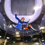 Two people in iFLY flight suits float mid-air with arms outstretched, smiling and experiencing the thrill of indoor skydiving adventure.