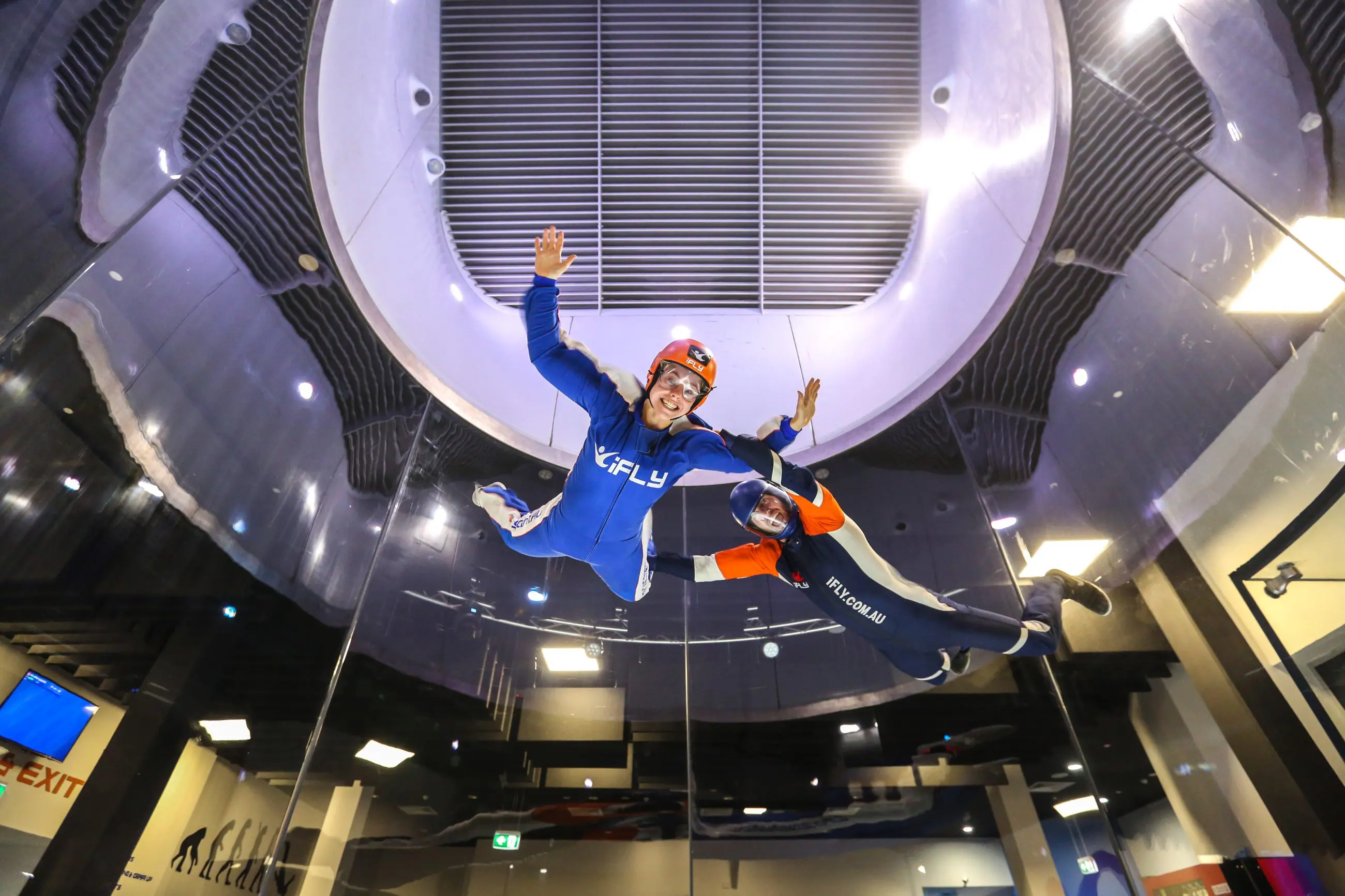 Two people in iFLY flight suits float mid-air with arms outstretched, smiling and experiencing the thrill of indoor skydiving adventure.