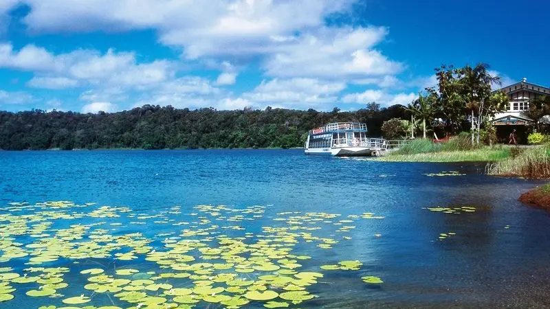Serene northern lake with vibrant lily pads, a quaint boat by Paronella Park’s scenic shore beneath a picturesque blue, cloudy sky.