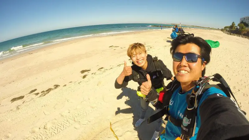 Two smiling skydivers in full kit give thumbs up after a Sunshine Coast Beach tandem skydive with blue skies and sea views.