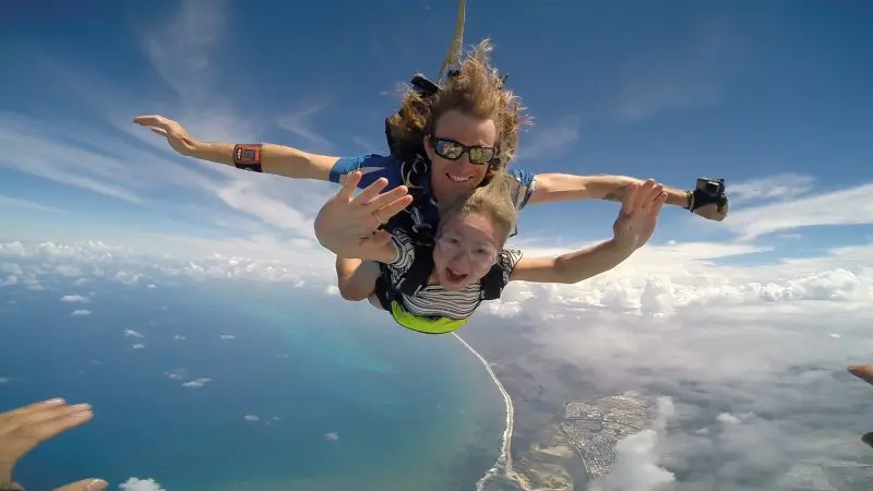 Adventurous tandem skydivers soar above Sunshine Coast Beach, smiling and waving over sparkling ocean waves, blue sky and clouds.