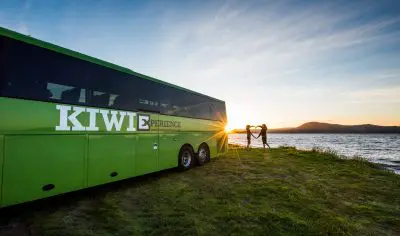 KIWI EXPERIENCE green coach parked by a scenic lake at sunset, two travellers stand by the water enjoying vibrant New Zealand views.