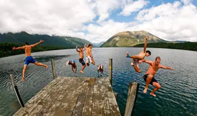 Group of seven jumps from a wooden jetty into a scenic mountain lake, enjoying the Grand Kiwi Experience on a cloudy day in New Zealand.