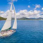Experience the Spank Me 2 Day 2 Night Whitsunday Sailing trip with a white yacht gliding by lush green hills beneath dramatic clouds.