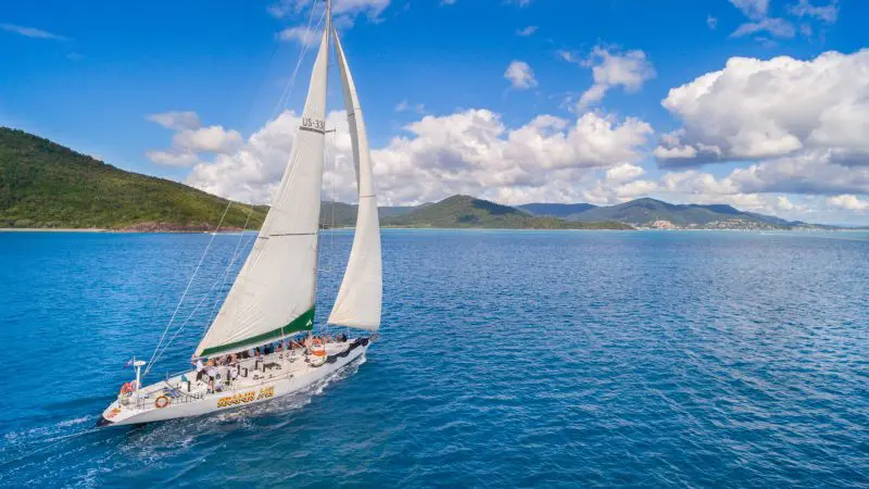 Experience the Spank Me 2 Day 2 Night Whitsunday Sailing trip with a white yacht gliding by lush green hills beneath dramatic clouds.