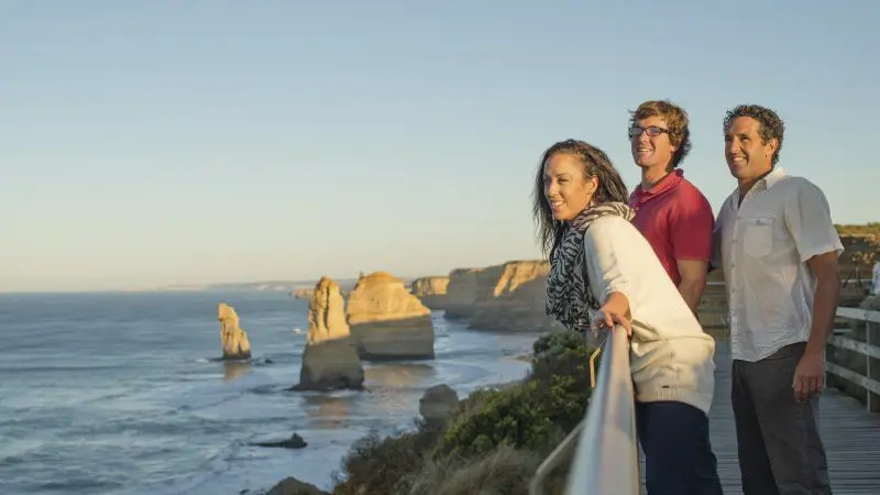 Three people enjoy a scenic sunset on a boardwalk during a 1 Day Great Ocean Road Sunset Tour with Go West Tours, breathtaking views.
