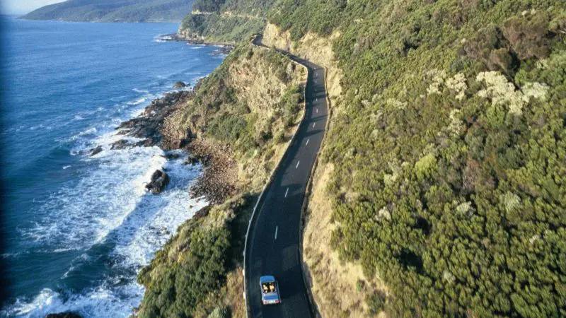 Experience the iconic 1 Day Great Ocean Road 12 Apostles Tour with Go West Tours as a car winds between lush green hills and the stunning sea.