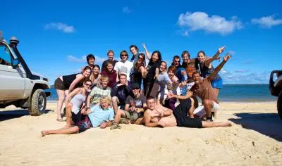 Group of happy travellers posing on a sunny East Coast Explorer tour, sandy beach, parked vehicles, and vibrant blue sky in background.