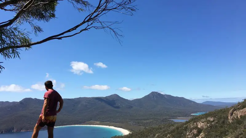 Explorer on a rocky ledge overlooks Wineglass Bay Tour, Richmond, forests, and mountains under bright clear skies—Tasmania adventure.