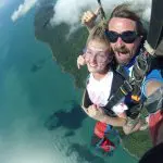 Tandem skydivers free-fall above scenic coastline, grinning at camera, with blue ocean waves and lush green landscape below.