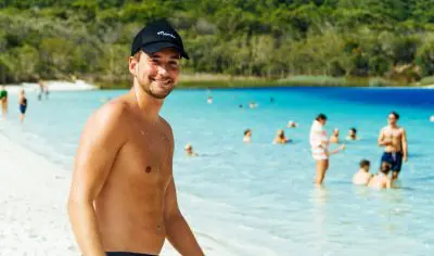 Happy man in swimming trunks enjoys a scenic beach on his 27-day tour, with crystal-clear blue water and swimmers in the background.