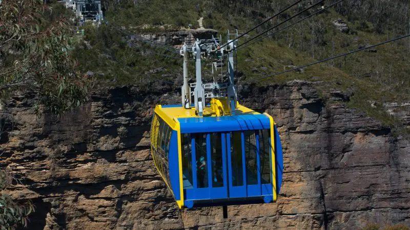 Experience breathtaking views as a blue and yellow cable car soars above a rugged gorge on the ultimate 1 Day Blue Mountains Tour.