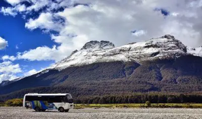 Tour coach on gravel beside Dart River, framed by majestic snow-capped mountains and dramatic blue sky with clouds in New Zealand.
