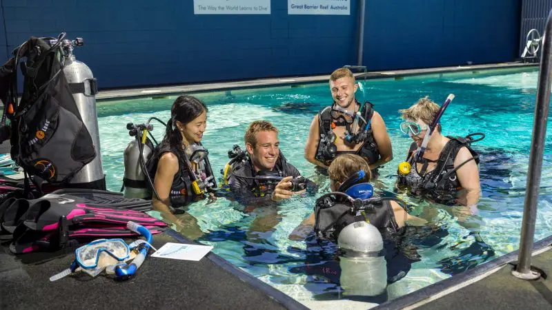 Five divers in full scuba gear smile and interact beside an indoor swimming pool during a 4 Day Learn to Dive course for beginners.