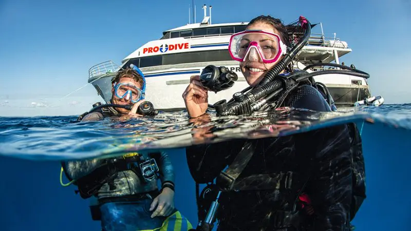 Two happy scuba divers beside a PRODIVE boat during a 4-day eLearning Liveaboard, enjoying crystal-clear blue skies on the water.