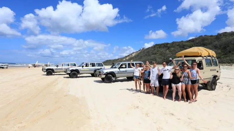 Adventure seekers stand next to rugged off-road vehicles on a sandy beach beneath a vibrant blue sky with fluffy white clouds.
